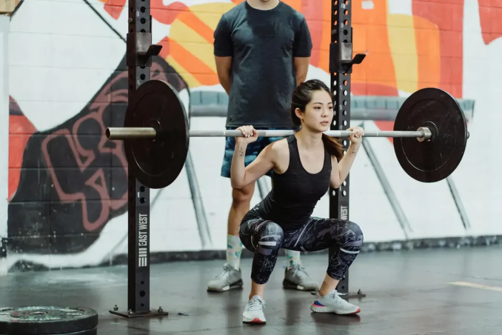 Woman performing a barbell back squat in a gym while a coach stands nearby for support.