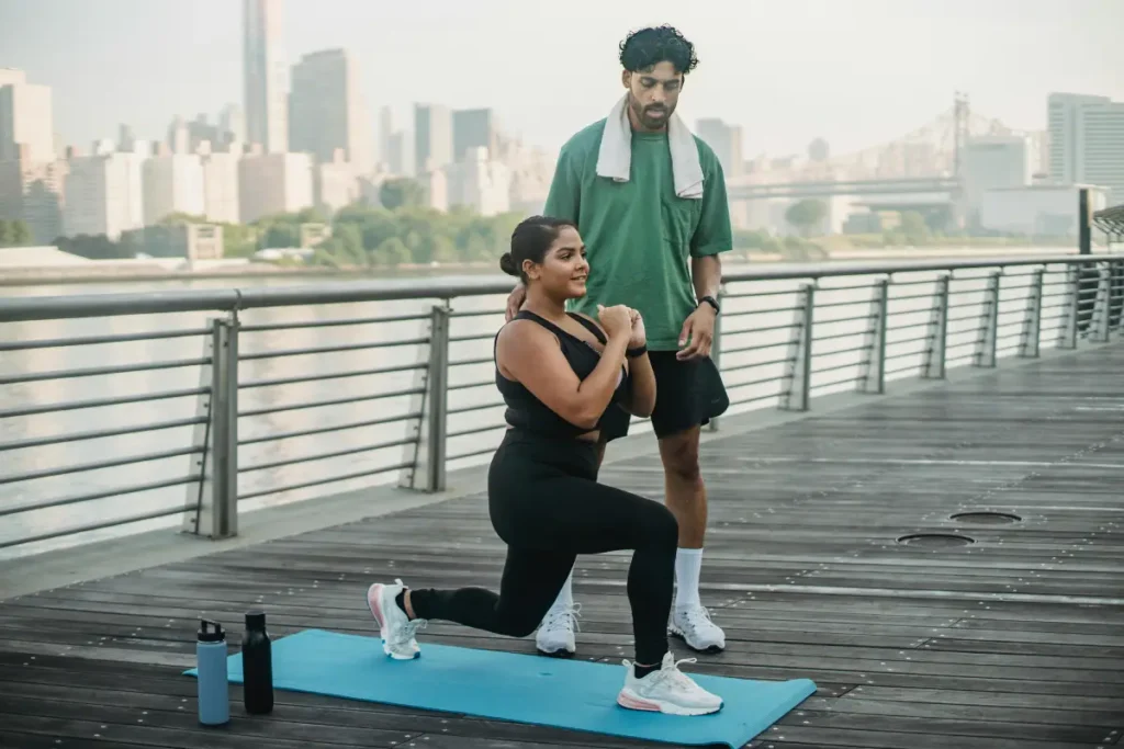 Woman doing a lunge exercise on a yoga mat outdoors with a fitness coach supervising her.