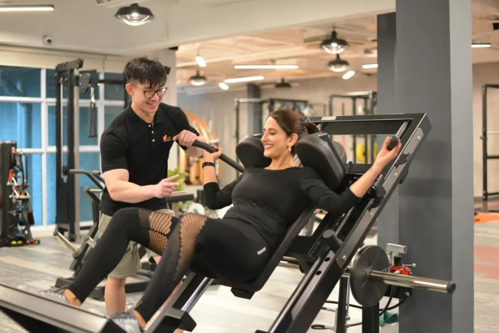 Woman performing hack squats with personal trainer assistance in a modern gym